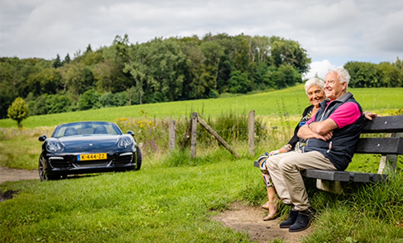 Jan en Miep zitten op een houten bankje in het groen met op de achtergrond hun Porsche Boxster