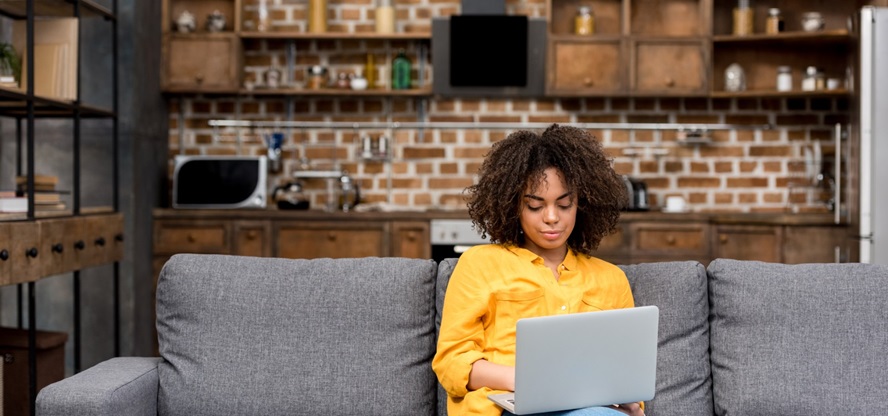 Een vrouw in een gele bloes zit op haar grijze bank met een laptop op schoot