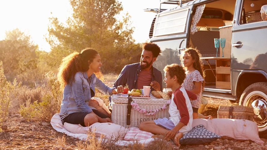 een gezin van 4 zit in het zonnetje op een picknickkleed naast een busje